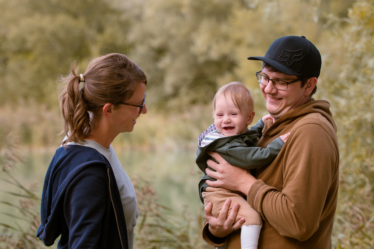 Mama und Papa stehen im Grünen und lachen ihr fröhliches Baby an, das Papa im Arm hält.